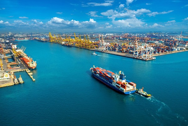 Beladenes Containerschiff von oben beim Einfahren in einen Containerhafen mit Kränen.